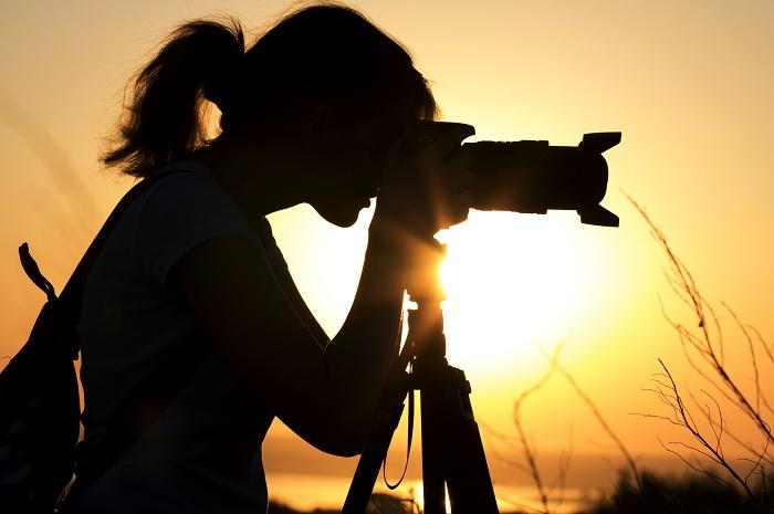 female photographer using camera on a tripod silhouetted by a setting sun with orange white glow